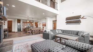 A spacious living area with a grey sofa, patterned ottoman, and a coffee table. A kitchen with wooden cabinets and a dining area with a wooden table and chairs are visible.