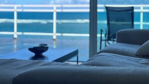 Una habitación moderna con un ventanal grande que da a un balcón y al mar. Hay un sofá y una mesita con un adorno encima.