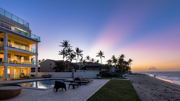 A beachfront hotel with a swimming pool, lounge chairs, and palm trees.