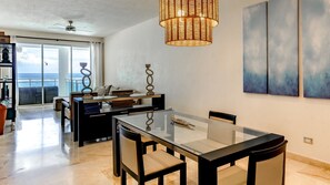 A modern dining area with a glass table and chairs, a wicker chandelier, and a view of the ocean through large windows. - Oceanfront Hot Tub Pool King Size Bed 305 (Kite Beach)