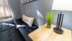 A modern room with a black leather sofa, a wooden side table with a lamp, a small potted plant, and a cushion with the word "Sunshine".