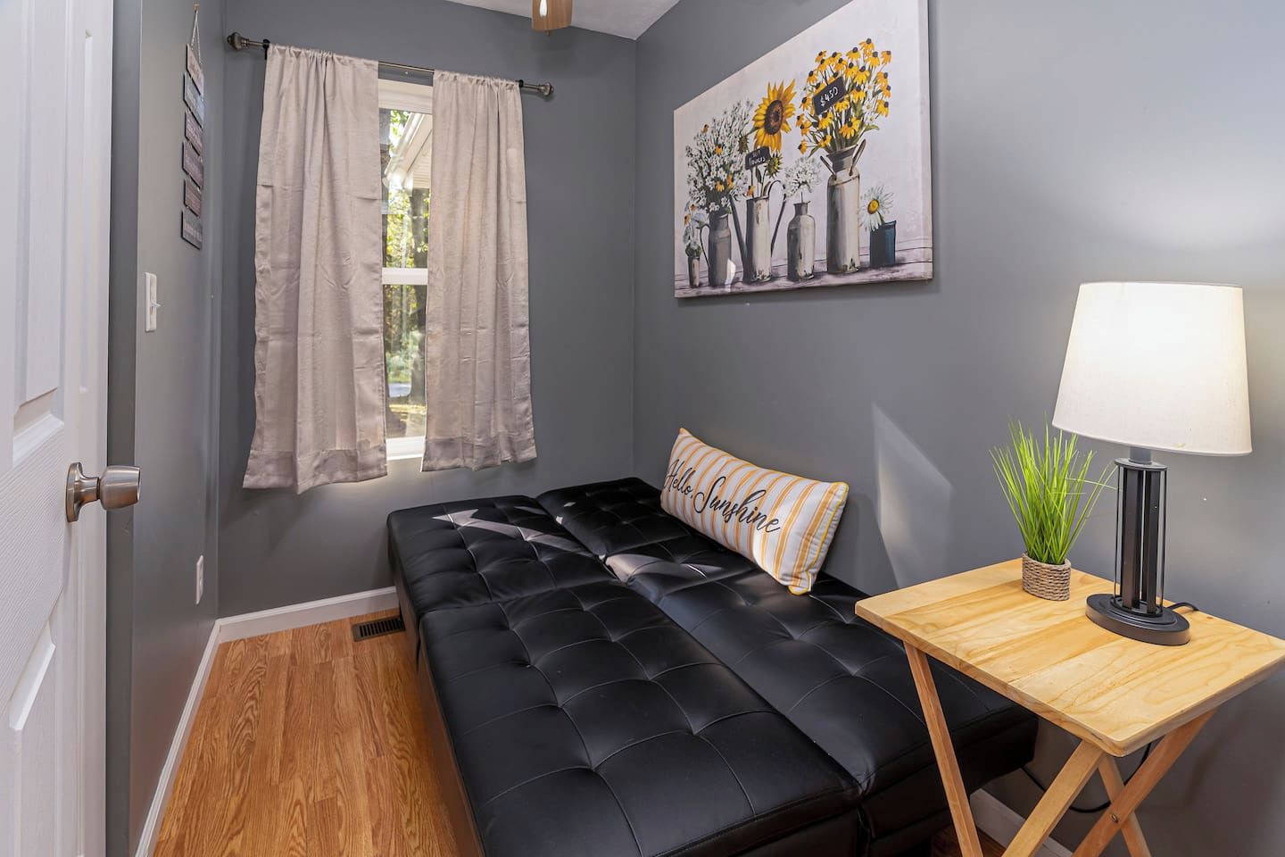 A room with a black leather sofa, a wooden side table with a lamp, a window with curtains, and a painting on the wall.