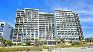 Un hotel de varios pisos con numerosos balcones, situado cerca de una playa de arena con palmeras.