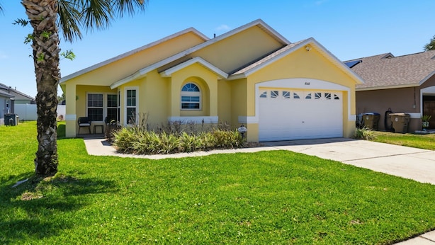 A yellow single-story house with a garage, a front lawn, and a palm tree.