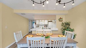 Un coin repas avec une table en bois et des chaises blanches, une cuisine en arrière-plan et un luminaire au plafond.
