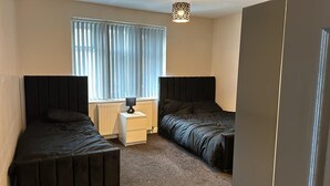 Une chambre avec deux lits simples, une table de chevet avec une lampe, un radiateur et une fenêtre avec des stores.