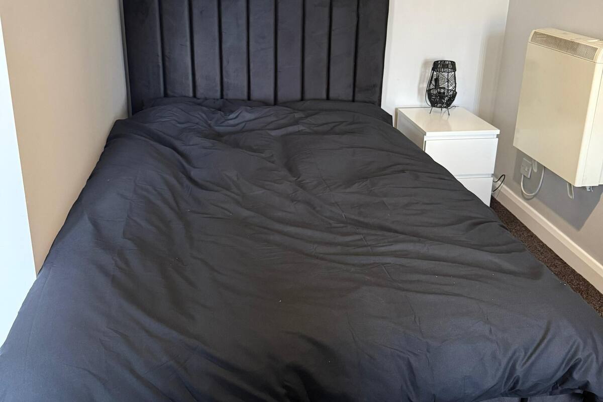 A bed with a black headboard and footboard, a white nightstand with a lamp, and a wall-mounted air conditioner.