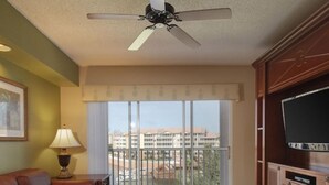 A living room with a brown leather sofa, a wooden coffee table, a TV on a wooden cabinet, a ceiling fan, and a balcony with a view of buildings. - Luxury Resort Minutes from Disney  (Kissimmee)