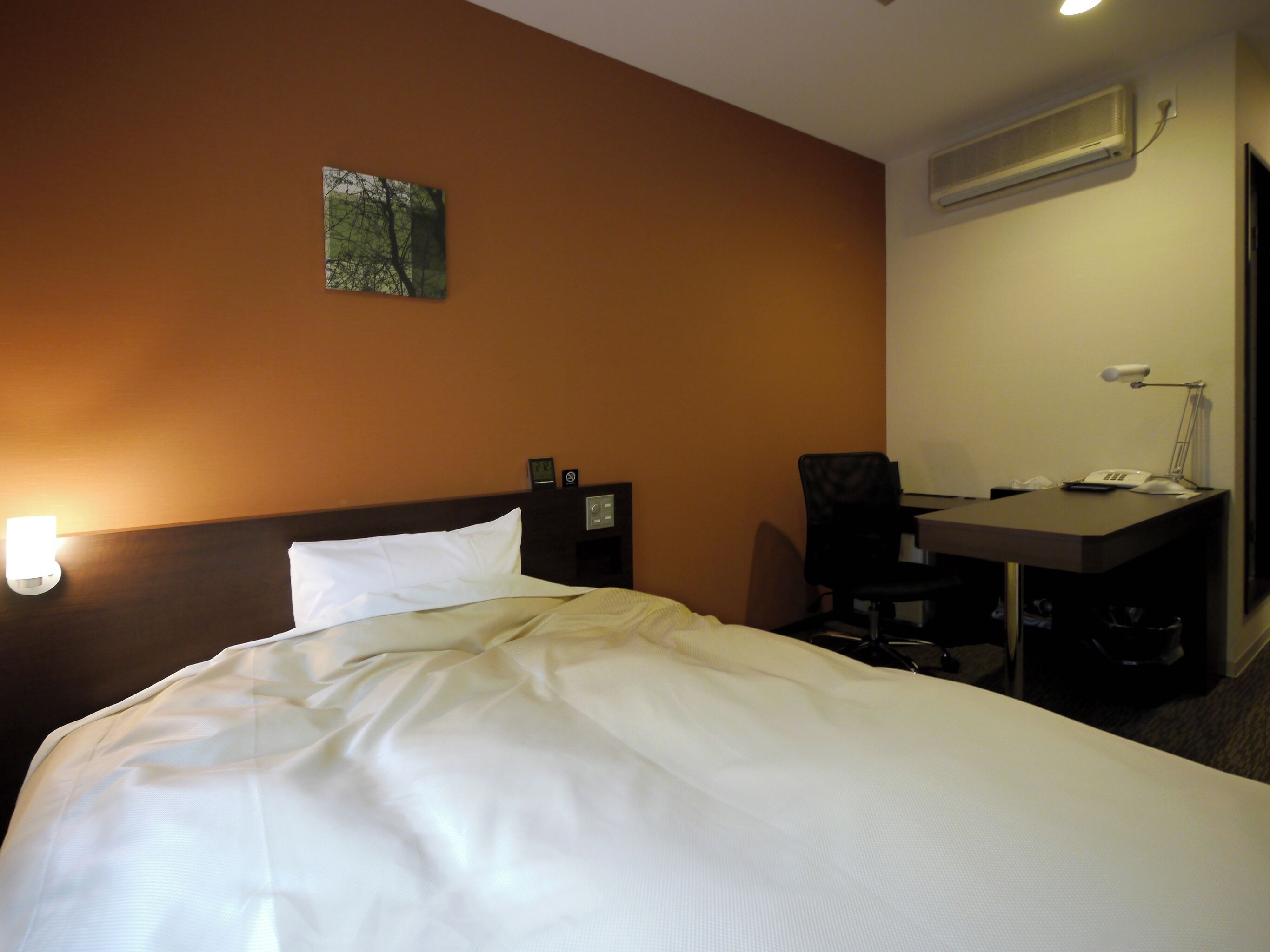 A hotel room with a bed, a desk, a chair, a nightstand, a wall-mounted air conditioner, and a small painting.