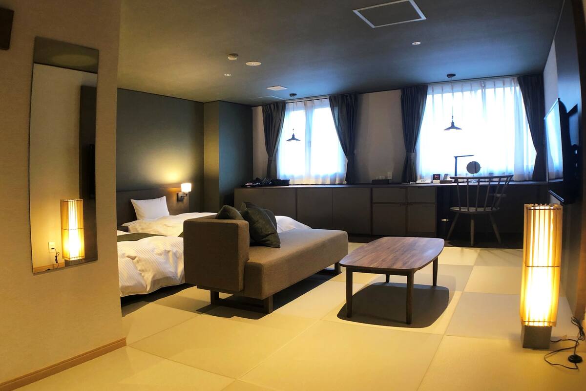 Een moderne hotelkamer met een bed, een bank en een kleine tafel. Er zijn twee ramen met gordijnen, een bureau en een stoel.