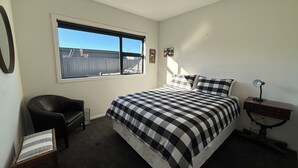 A bedroom with a large bed, a chair, a desk, a window, and a small table with a lamp. - Pedal and Play - Cromwell Holiday Home (Cromwell)