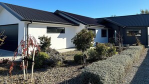 A modern two-story house with a sloped roof, a garage, and a well-maintained garden with various shrubs and a small tree. - Pedal and Play - Cromwell Holiday Home (Cromwell)