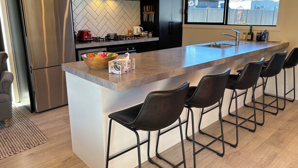 A modern kitchen with a large island, bar stools, a refrigerator, and a window with a mountain view. - Pedal and Play - Cromwell Holiday Home (Cromwell)