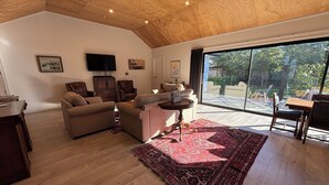 A spacious living room with a wooden ceiling, a large window, and a red patterned rug. - Pedal and Play - Cromwell Holiday Home (Cromwell)