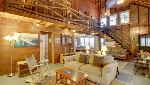 A spacious living room with a high ceiling, wooden beams, a staircase, and a large window. - Rustic Cliff-top Cabin at the Tip of Lake Champlain's Barney Point (Colchester)