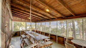 A rustic wooden cabin with a large dining table and benches, large windows offering a view of trees and a body of water. - Rustic Cliff-top Cabin at the Tip of Lake Champlain's Barney Point (Colchester)