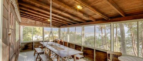 A rustic wooden cabin with a large dining table and benches, large windows offering a view of trees and a body of water.