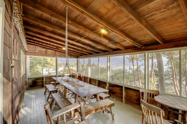 A rustic wooden cabin with a large dining table and benches, large windows offering a view of trees and a body of water.