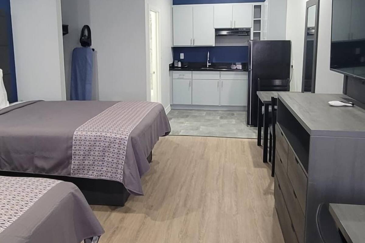 A modern hotel room with two beds, a kitchenette, a desk, and a TV.