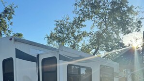 A white recreational vehicle with a 'Cugar by Regency' logo, parked on a gravel surface with trees in the background.