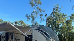 A large RV with a covered porch area, a wooden deck, and a gravel ground.