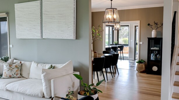 A modern living room with a white sofa, dining area with chairs, a chandelier, and a staircase.