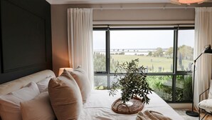 A bedroom with a large window, a bed with beige bedding, a bedside lamp, and a potted plant.