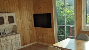 A dining area with a wooden table and chairs, a television mounted on the wall, and a window with a view of greenery. - Vacation home with outdoor sauna in the Thuringian Forest (Schleusegrund)