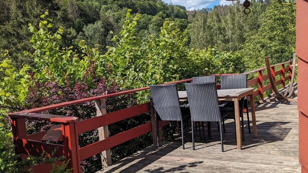 A wooden deck with a red railing, a table with chairs, and a bell in the background. - Vacation home with outdoor sauna in the Thuringian Forest (Schleusegrund)