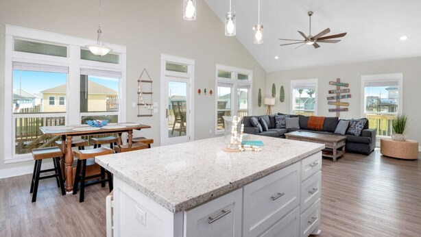 A spacious kitchen with a large island, a dining area with wooden chairs, a grey sofa, and a ceiling fan. - 3 Bdr/3 Bth, King Bed, No Fees (Bolivar Peninsula)