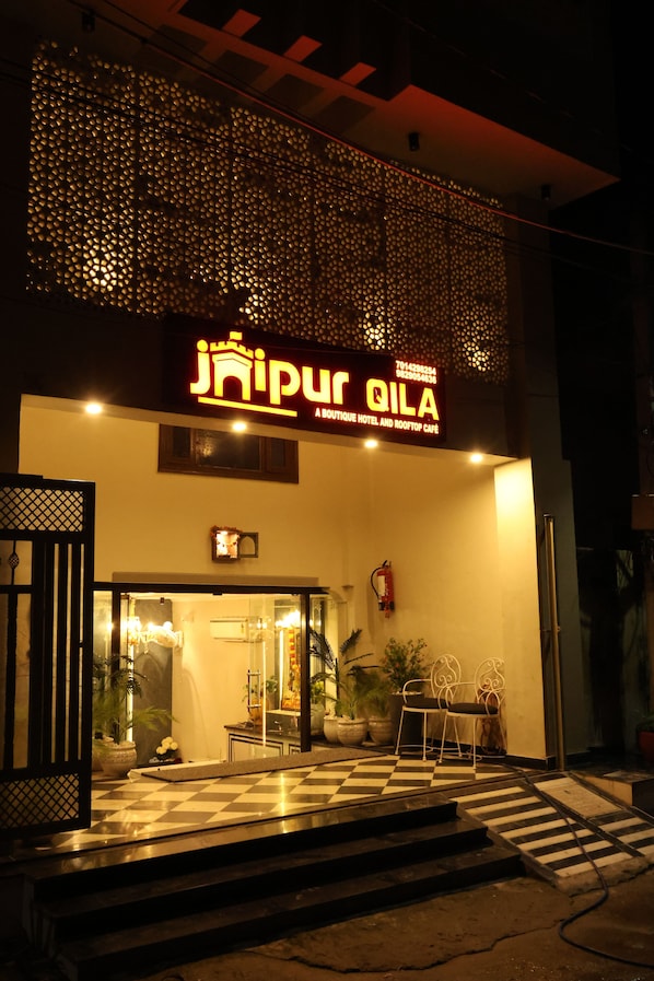 A hotel entrance with a checkered floor, potted plants, and a sign reading "Jai Pur Qila Boutique Hotel and Rooftop Cafe".