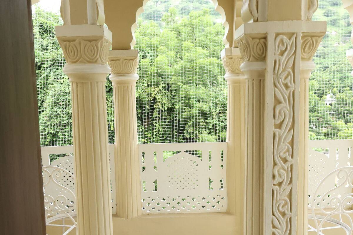 Un couloir orné de colonnes et surmonté d’une arche décorative, qui mène à un balcon offrant une vue sur la verdure.