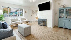A spacious living room with a fireplace, a television mounted on the wall, and a large window offering a view of the outside.