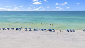 A beach with clear turquoise water and a sandy shore. There are several blue umbrellas and beach chairs set up along the beach.