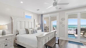 A bedroom with a large bed, a ceiling fan, a nightstand, and a view of the ocean through a sliding door.