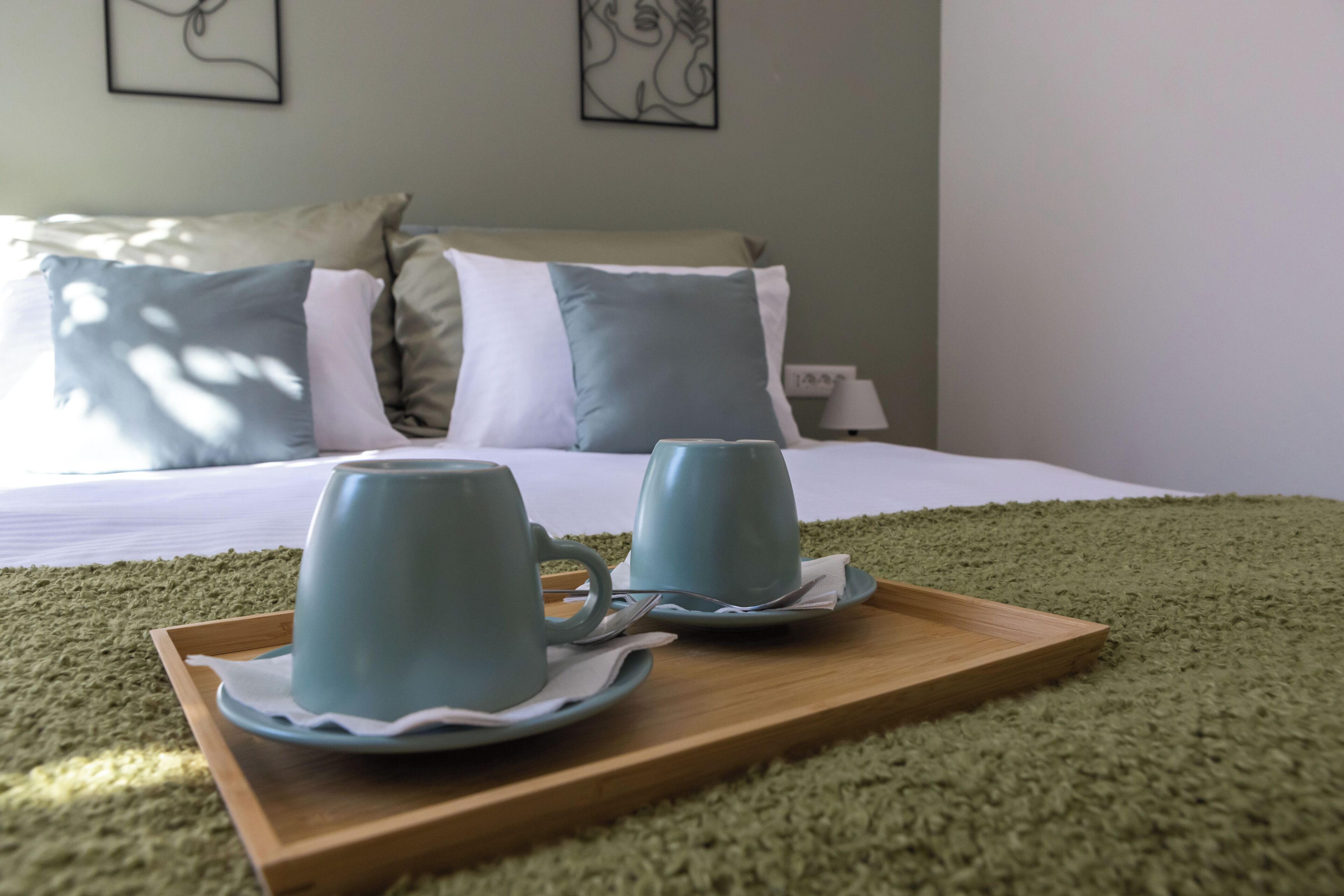 A neatly made bed with pillows and a tray with two cups and saucers on a green carpet.