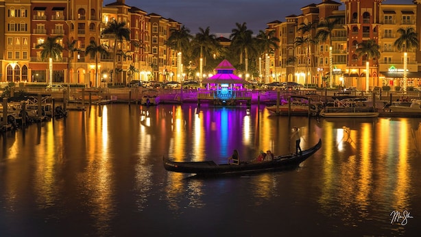 Un hotel frente al agua con varias construcciones, un quiosco central iluminado y un bote en el agua.