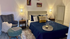 Un dormitorio con una cama, dos mesitas de noche, una silla, un pequeño otoman y decoración en la pared.
