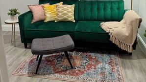 Un sofá verde con cojines estampados, un reposapiés gris, una alfombra decorativa y un letrero de neón en la pared.