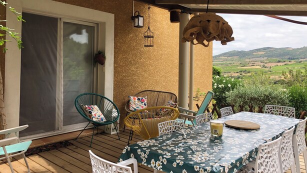 A covered outdoor patio with a table set for dining, surrounded by wicker chairs and a white chair with a colorful cushion.