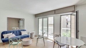A modern living room with a blue sofa, a round coffee table, and a dining table with chairs. There is a large window with a view of the outside. - An Attractive flat near by St-Germain, Le Bon Marché (Paris)