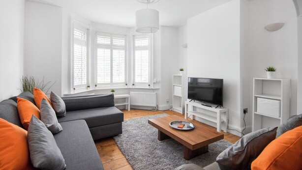 Un salon moderne avec un canapé gris, des coussins orange, une table basse en bois, un téléviseur posé sur un meuble blanc et une grande fenêtre avec des stores.