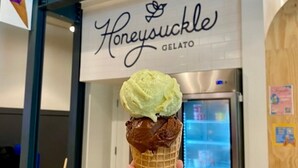 A hand holding a waffle cone with two scoops of ice cream in front of a store named 'Honeyyuckle Gelato'.
