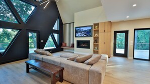 Ein modernes Wohnzimmer mit einer großen Eckcouch, einem Kamin, einem Fernseher und großen Fenstern mit Blick ins Freie.