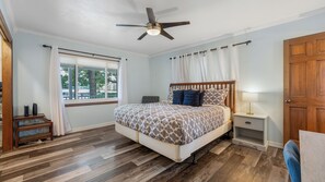 Ein Schlafzimmer mit einem Bett, einem Nachttisch, einem Deckenventilator, einem Fenster mit Vorhängen und einer Holztür.
