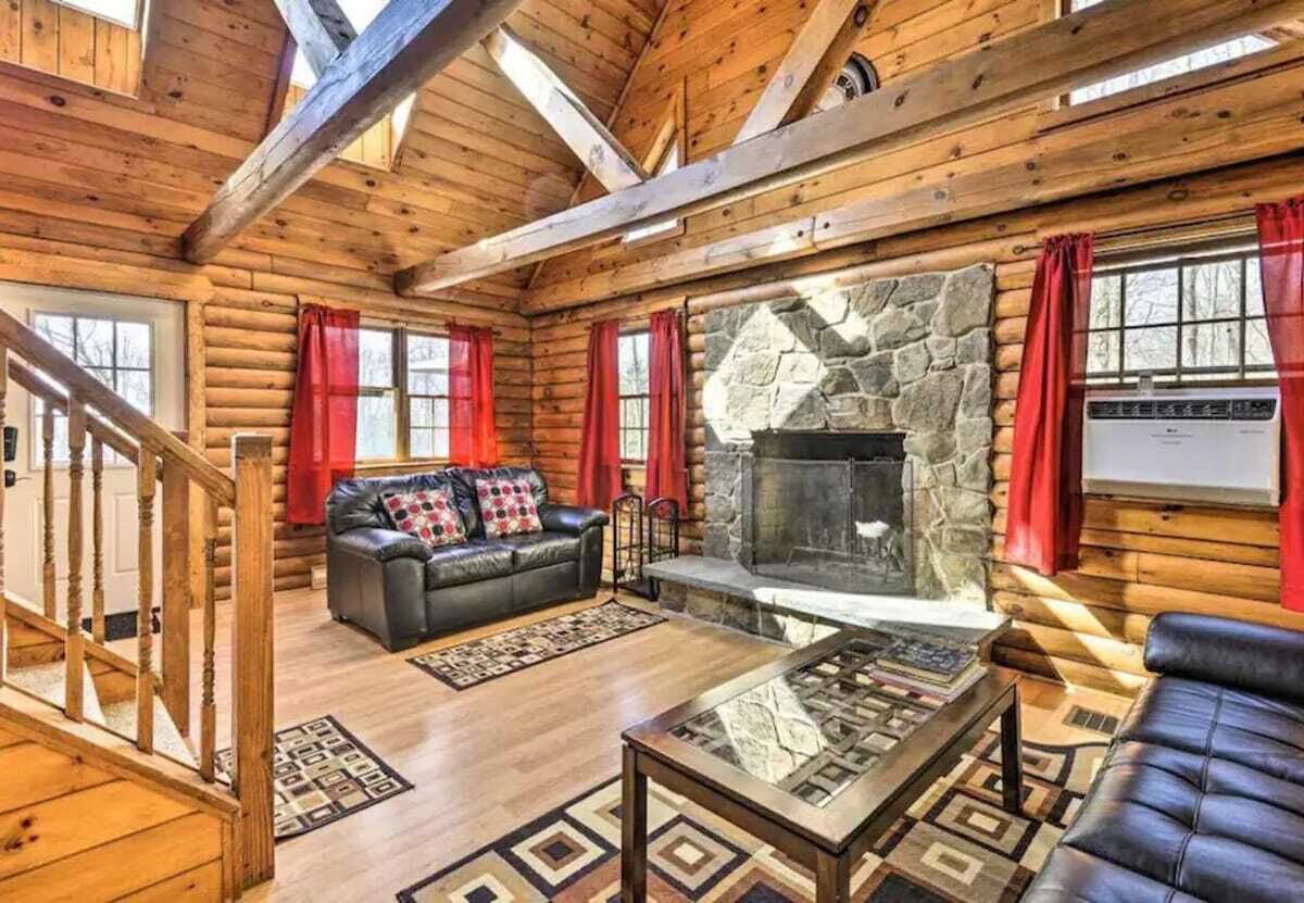 A log cabin interior with a stone fireplace, leather sofas, and a wooden staircase.