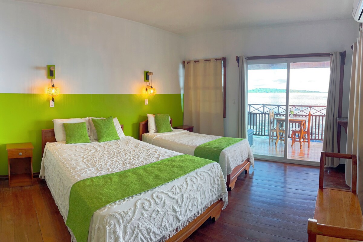 A hotel room with two beds, a balcony with a view of the sea, and a wooden floor.