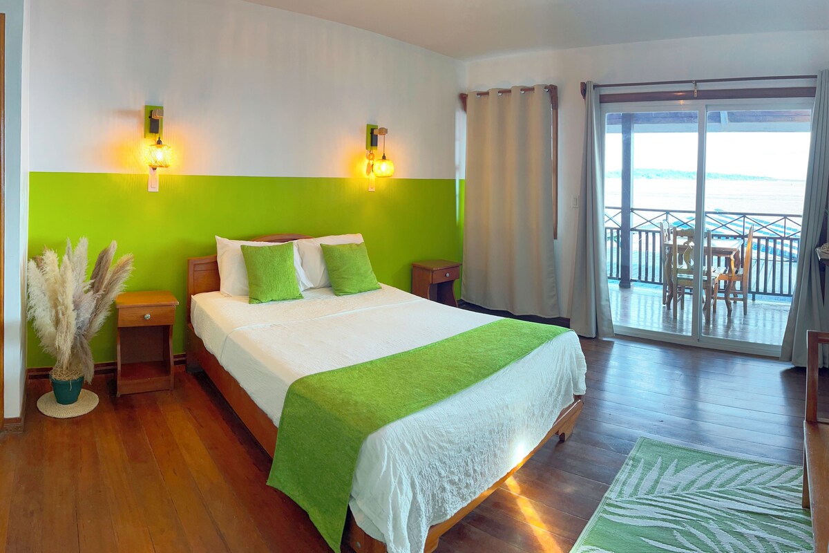 Un dormitorio con una cama grande, ropa de cama verde y blanca, dos lámparas de noche, una mesita de noche, una planta en maceta y vistas al balcón.