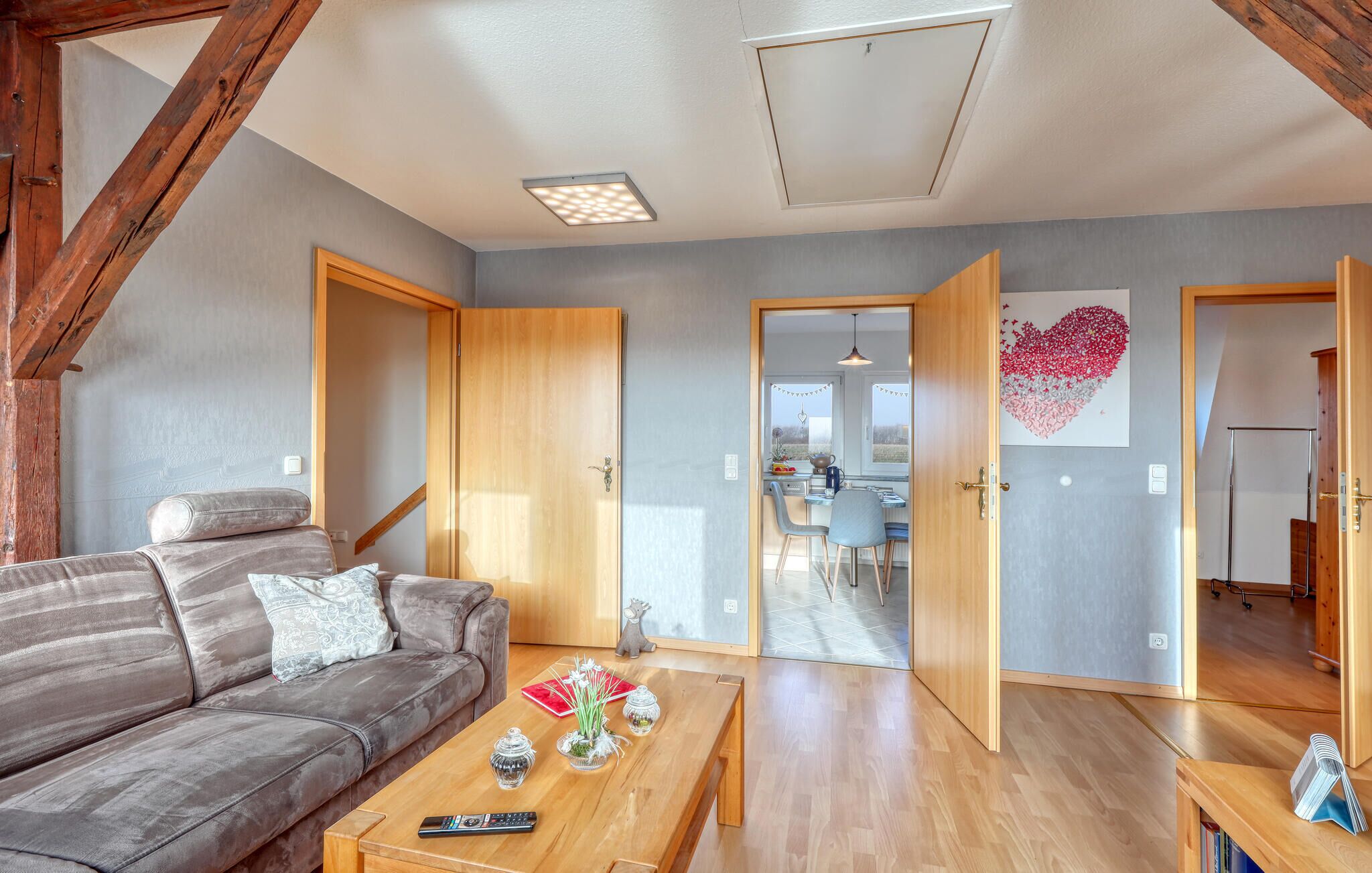 Een woonkamer met een bank, salontafel en afstandsbediening. Er zijn houten deuren en een gang die naar een keuken leidt.