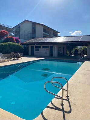 A swimming pool with a metal ladder, surrounded by a concrete deck. There are lounge chairs and a building with a solar panel roof in the background.